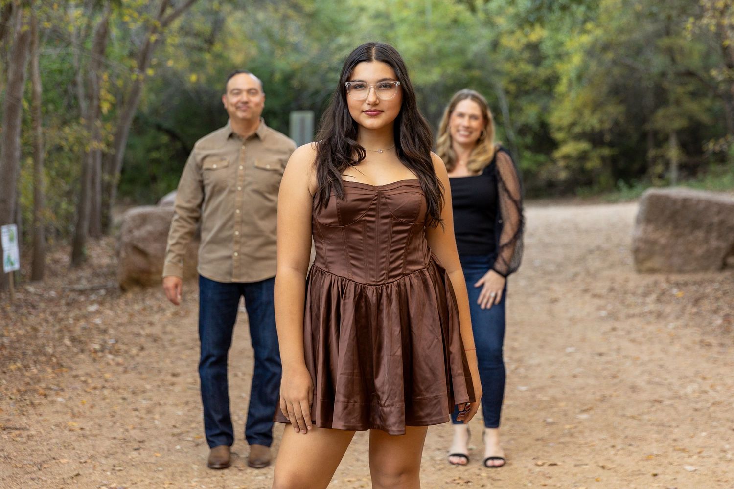 Three people pose on a dirt path in a wooded area, with someone in a brown dress in the foreground.