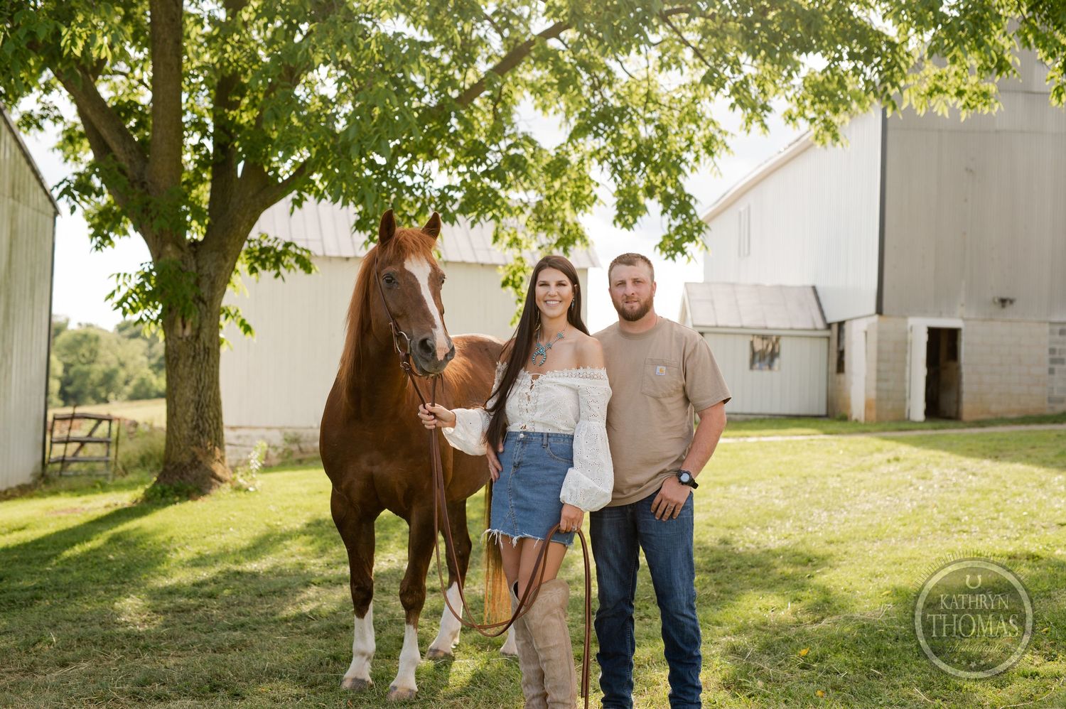 Family stands with brown and white horse under leafy tree near white barn.