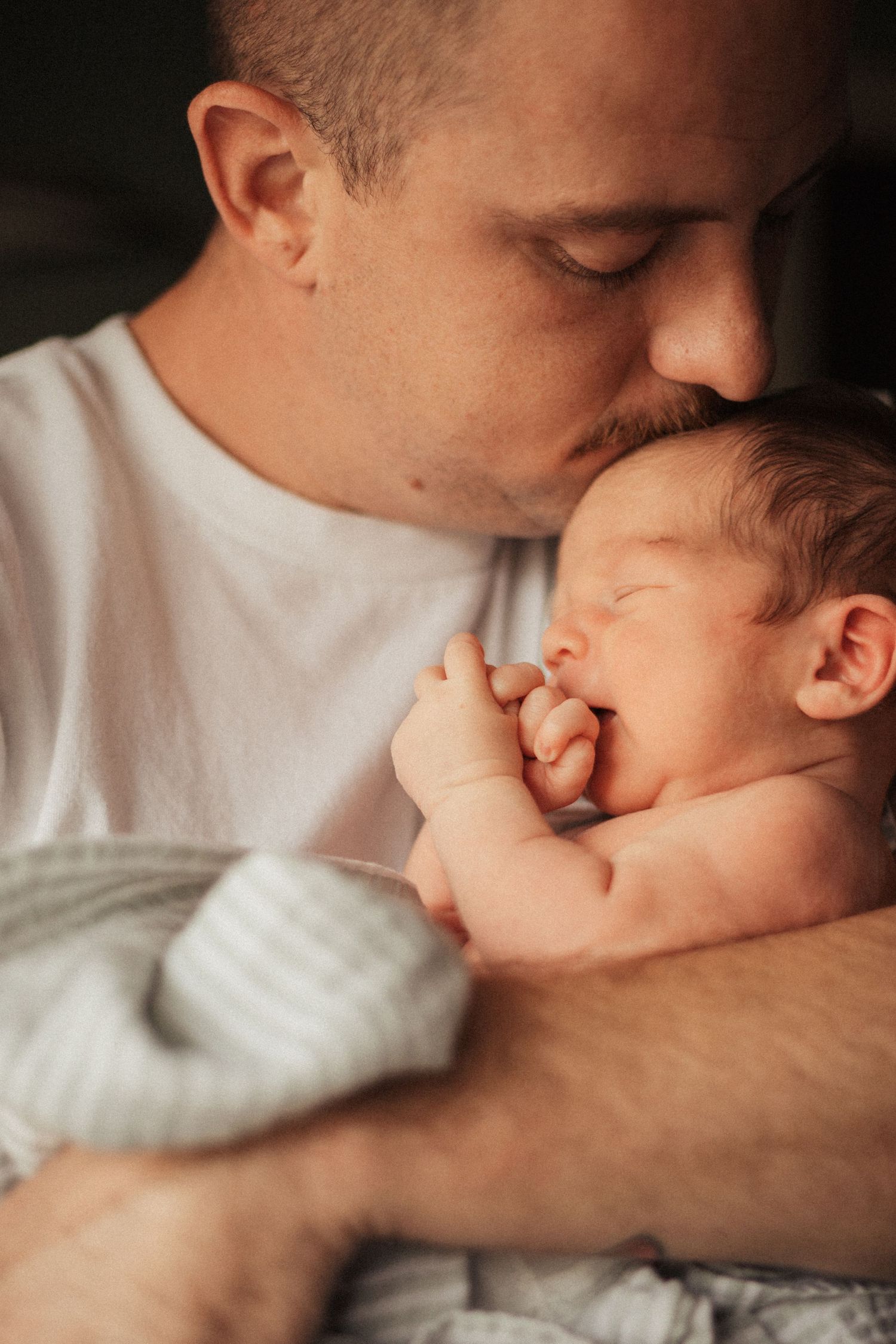 A tender parenting moment as an adult in a white shirt cradles and kisses a sleeping newborn baby.