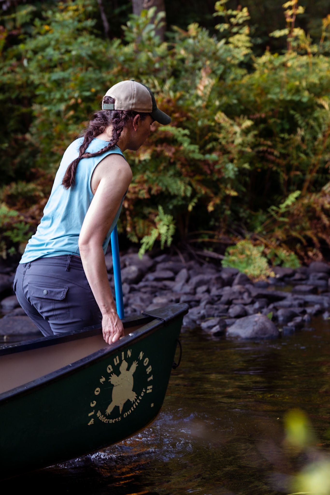 A paddler in a blue tank top sits at the edge of a green canoe on a calm river surrounded by forest scenery.
