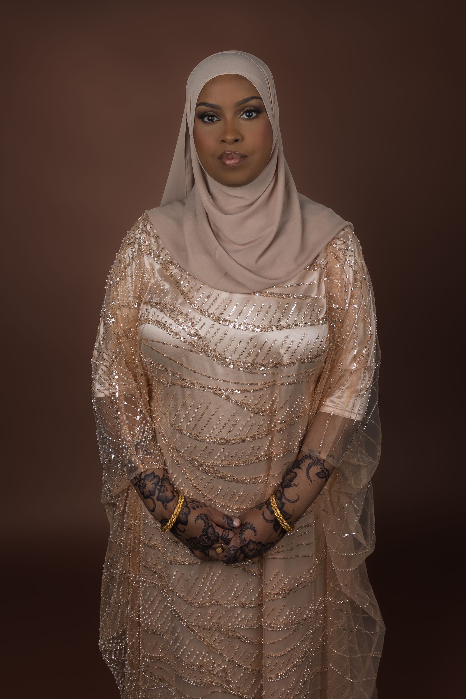 Beigeen iltapukuun ja yhteensopivaan hijabiin pukeutunut nainen poseeraa ruskeaa taustaa vasten valokuvastudiossa otetussa muotokuvassa.