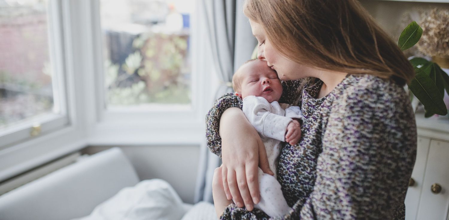 Mother holding sleeping newborn baby by window with soft natural lighting.