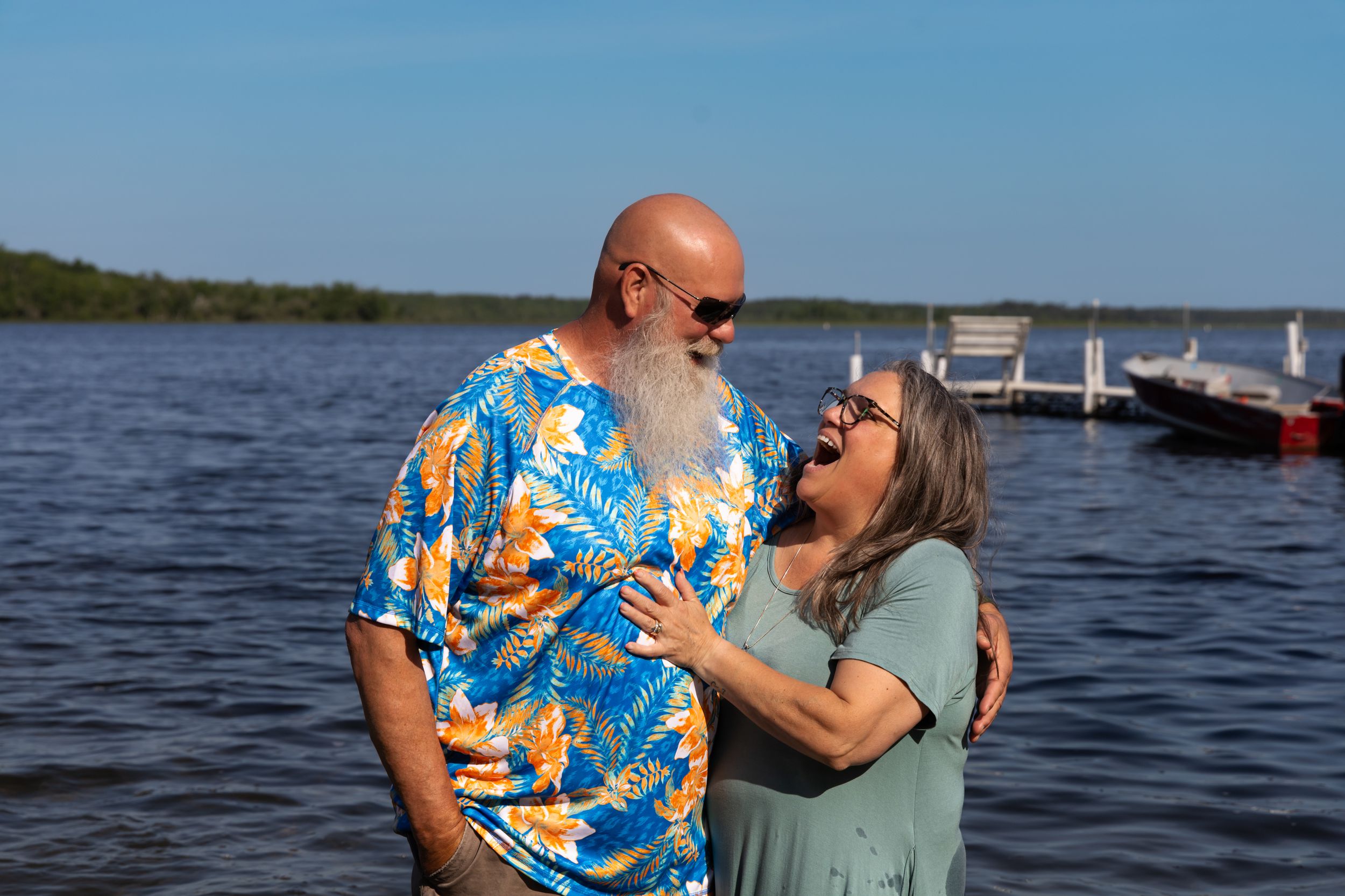 Affectionate couple embraces by a scenic lake on a sunny day with bright blue sky and calm waters.