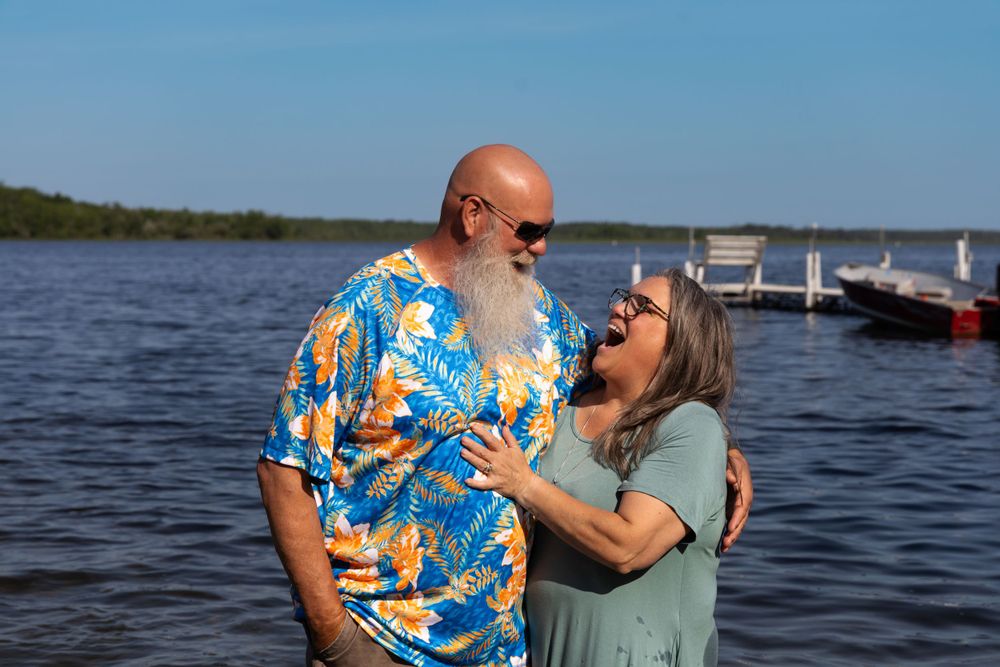 Affectionate couple embraces by a scenic lake on a sunny day with bright blue sky and calm waters.