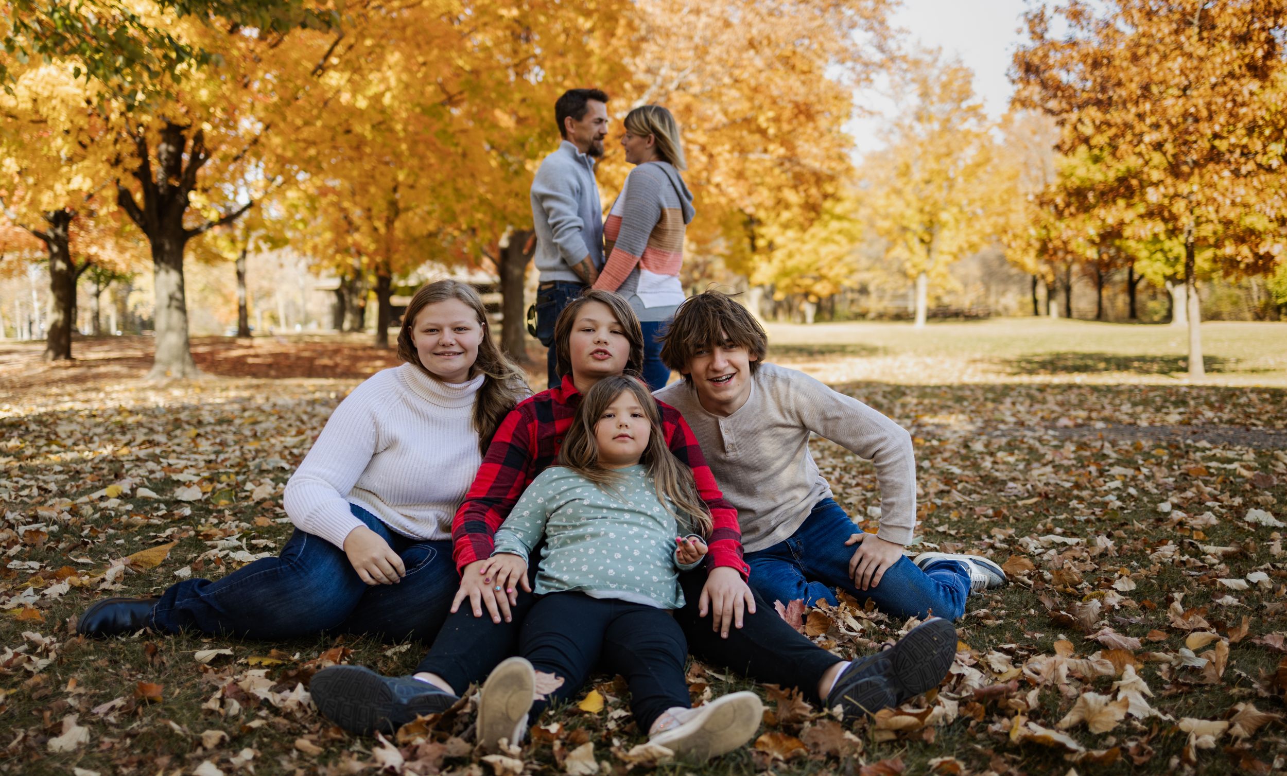 Happy family of five sitting close together on fallen autumn leaves in park.