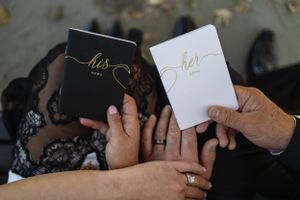Hands holding matching 'his' and 'hers' wedding vow books with gold lettering against a dark background.