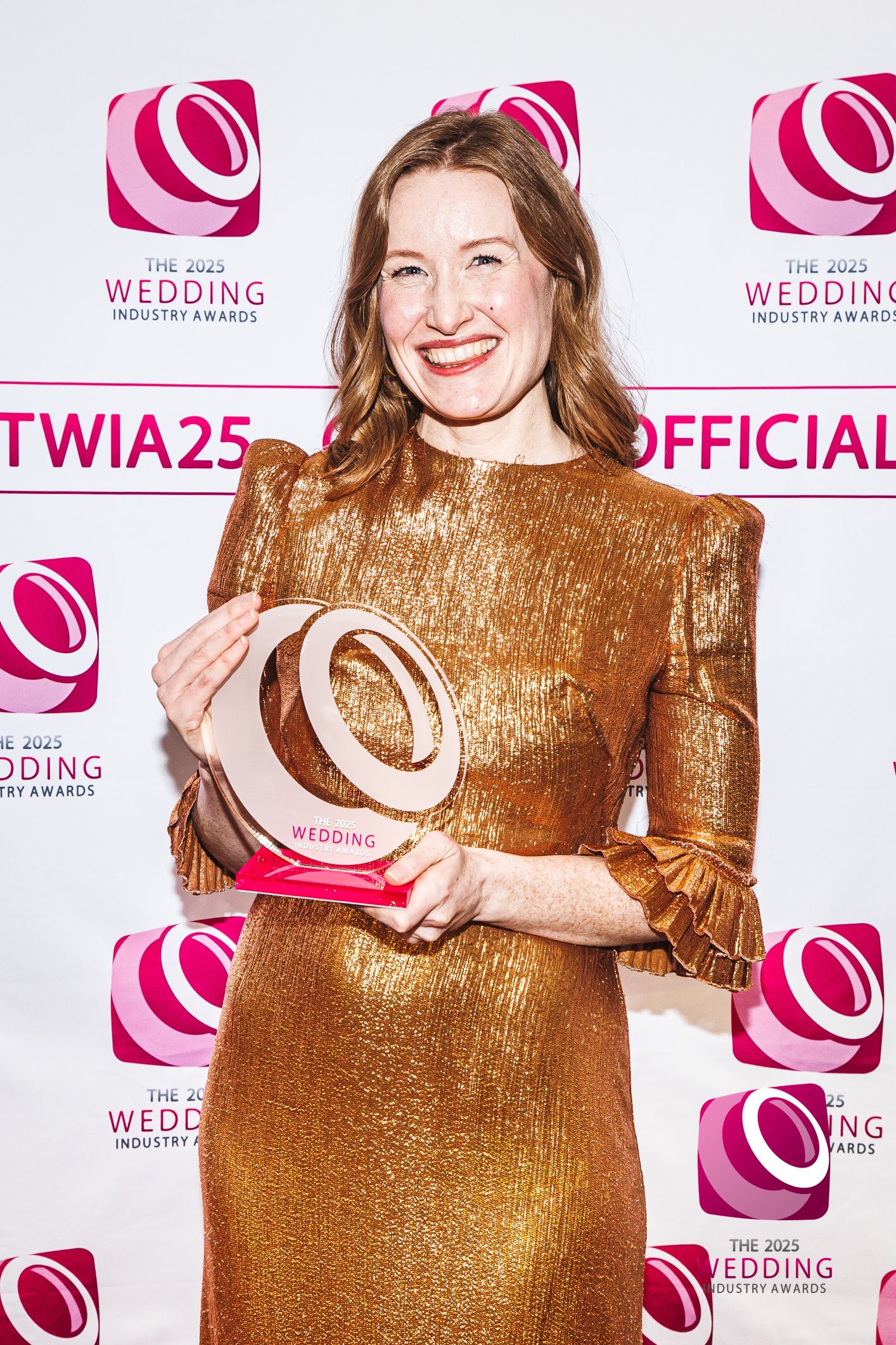 A smiling woman in a gold sequin dress proudly holds a Wedding Industry Awards trophy at the TWIA 2025 ceremony.