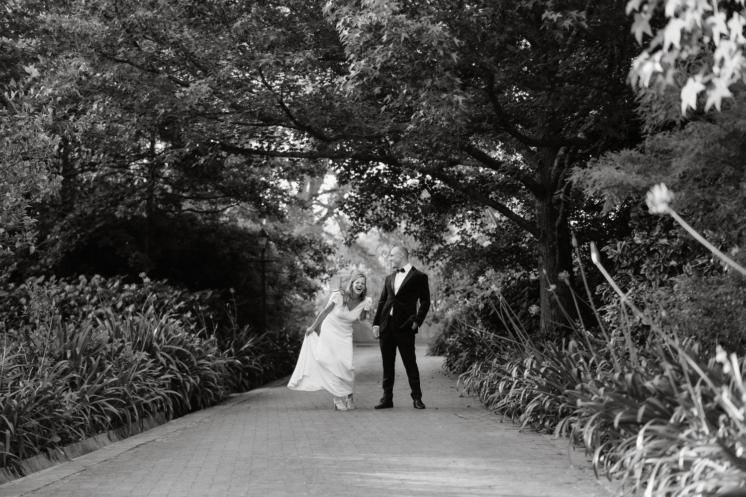 A black and white photo of a couple walking down a garden path surrounded by lush foliage.