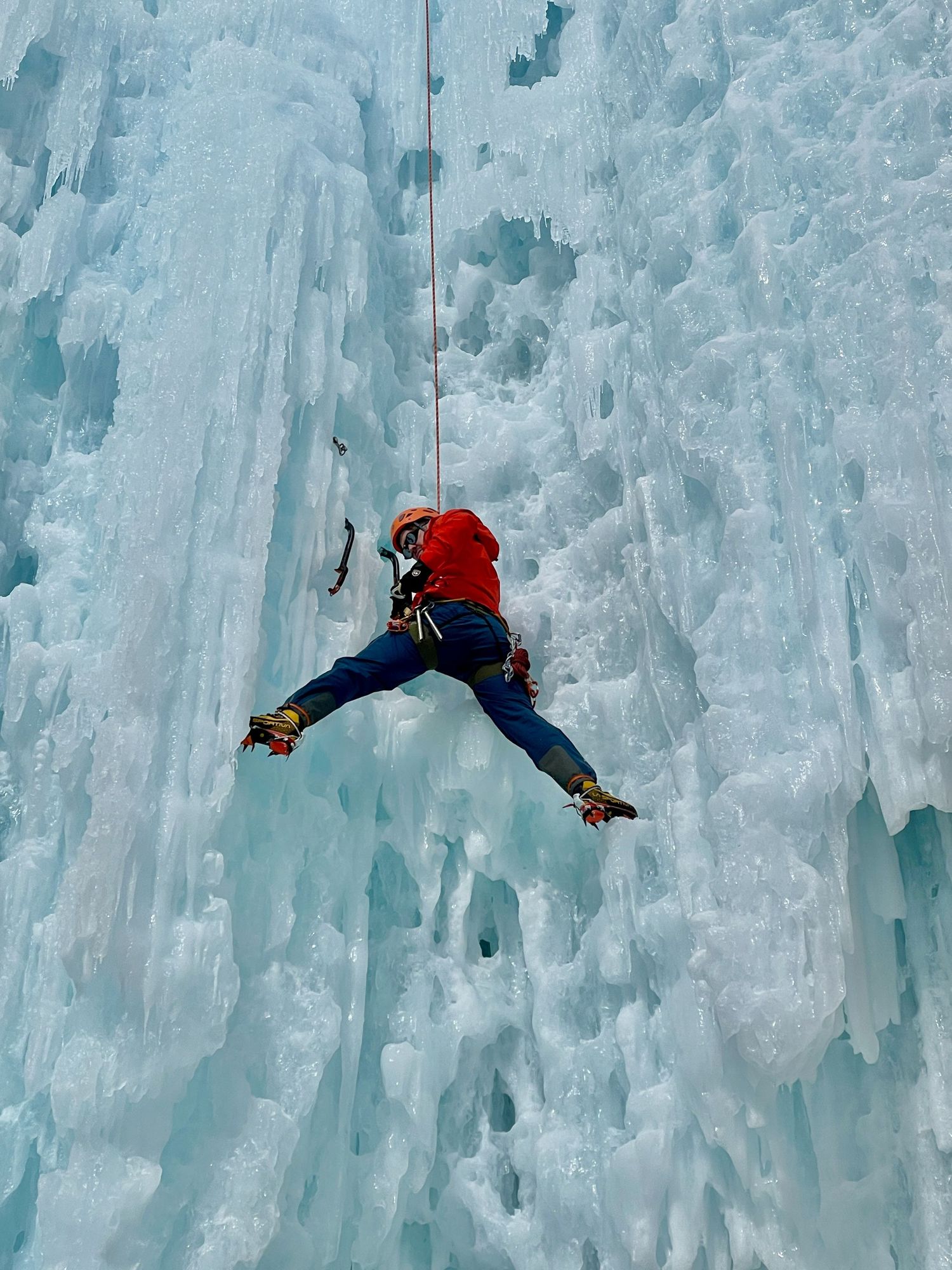 Evan Will Ice climbing in a red Arc'teryx jacket and blue Fjallraven pants climbs a towering frozen waterfall wall with climbing gear.
