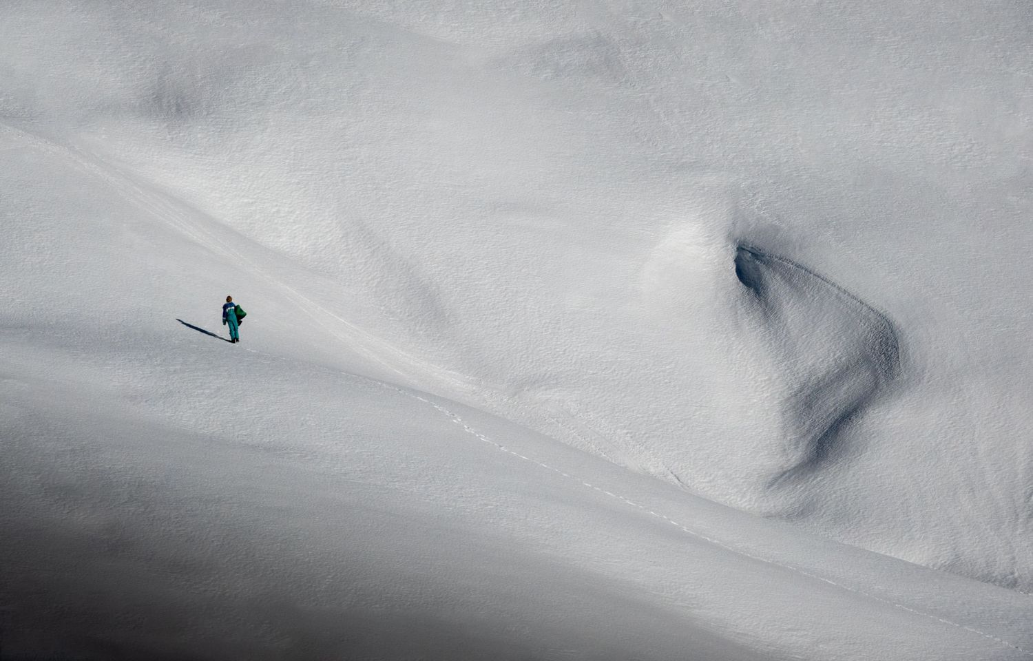 A tiny skier glides across vast white snow-covered mountain slopes under an overcast sky.