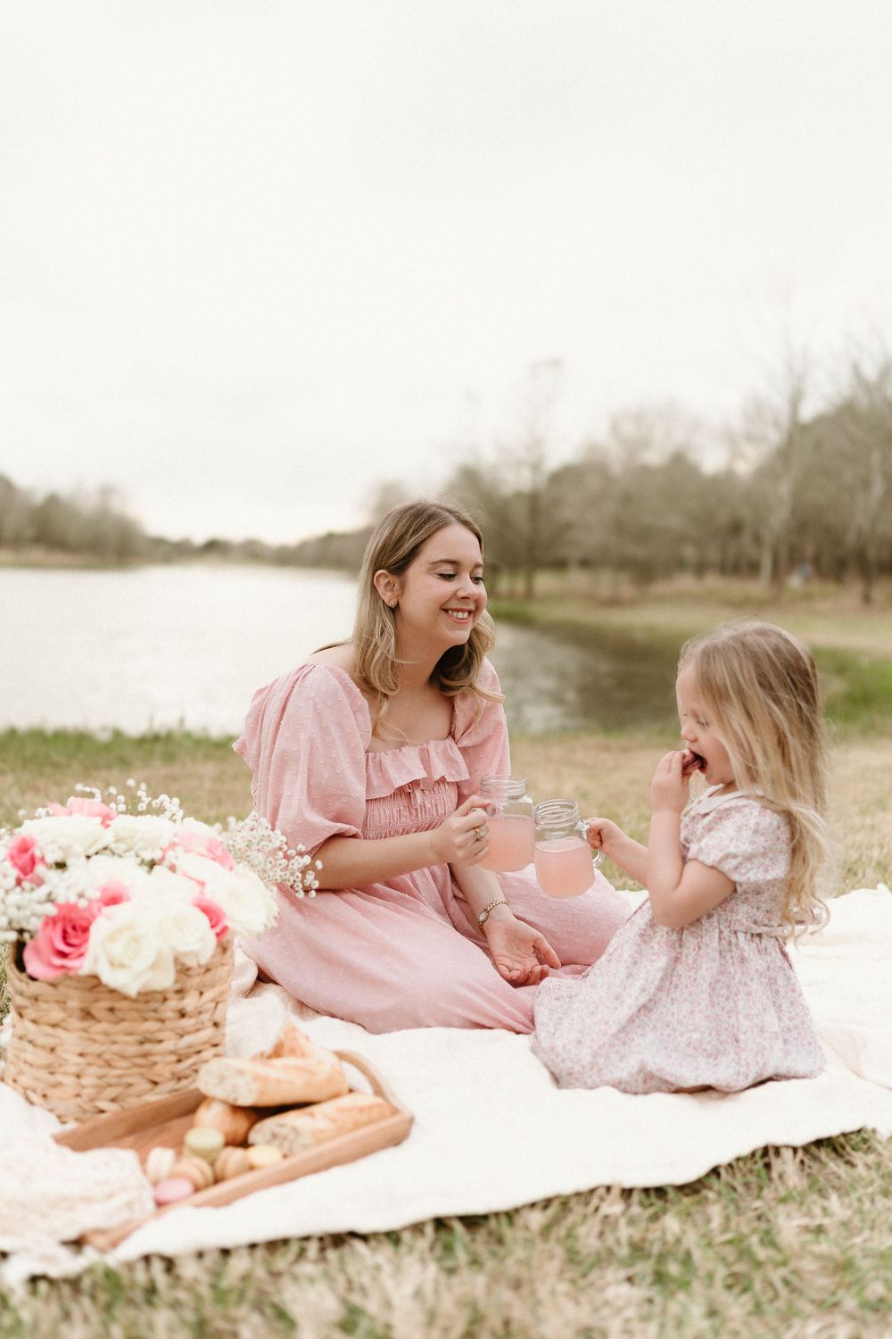 Picnic scene with pink dresses, roses, and wicker basket by the lake.