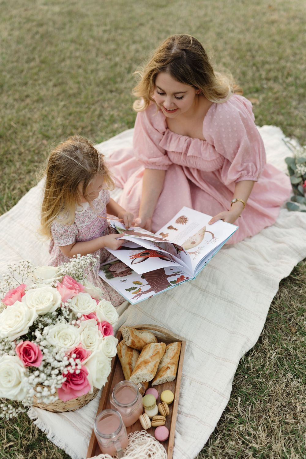 Mother and daughter share intimate reading moment on picnic blanket with flowers and treats.