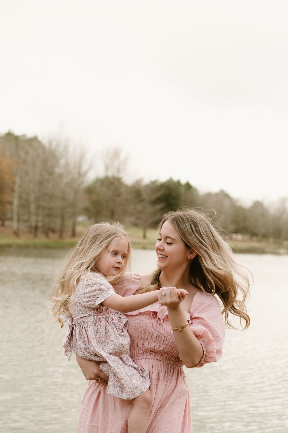 Two people in pink dresses share a joyful moment by a lake on a sunny afternoon.