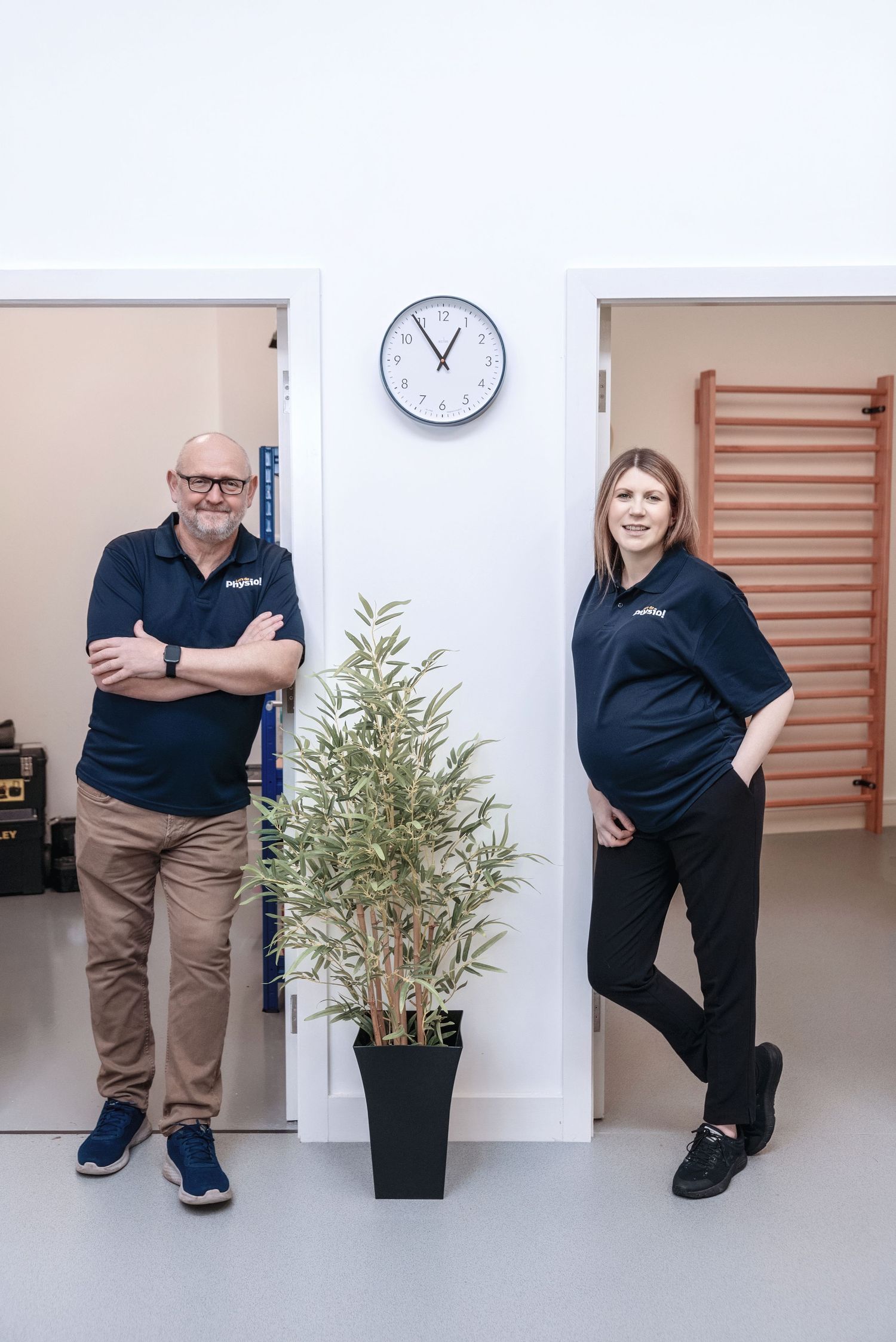 Two employees in navy uniforms stand casually against a white wall with a clock and potted plant between them.