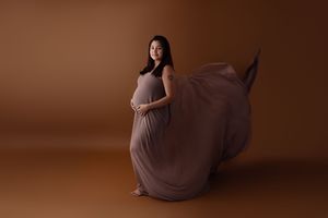 Pregnant woman in draped mauve gown sits elegantly on floor during studio maternity session.