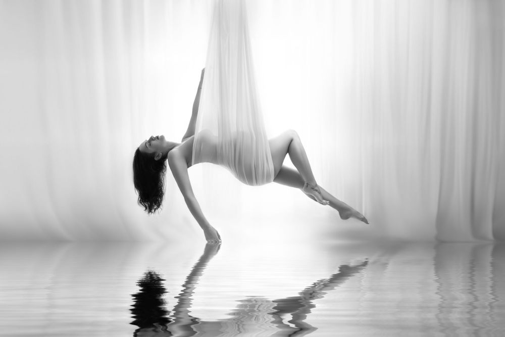 Artistic black and white photograph of a graceful dancer suspended in silk fabric with reflection on water surface.
