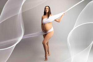 Artistic maternity photo with flowing white fabric against gray background showing elegant dance-like pose.