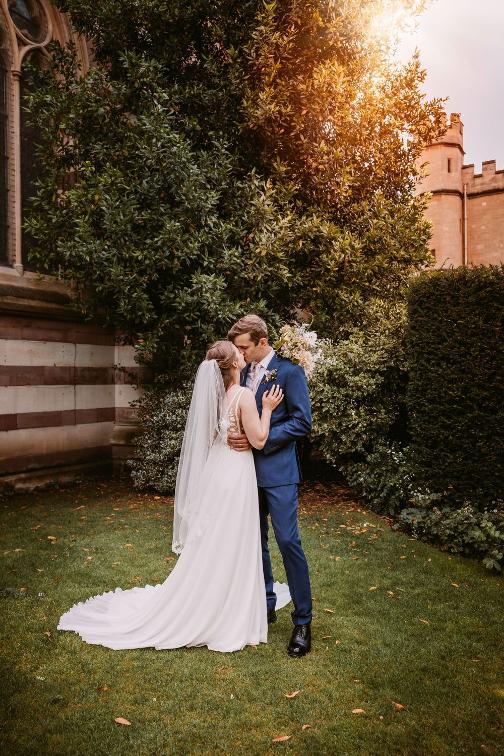 A romantic wedding portrait shows a couple embracing near a castle wall and autumn foliage at Balliol College, Oxford