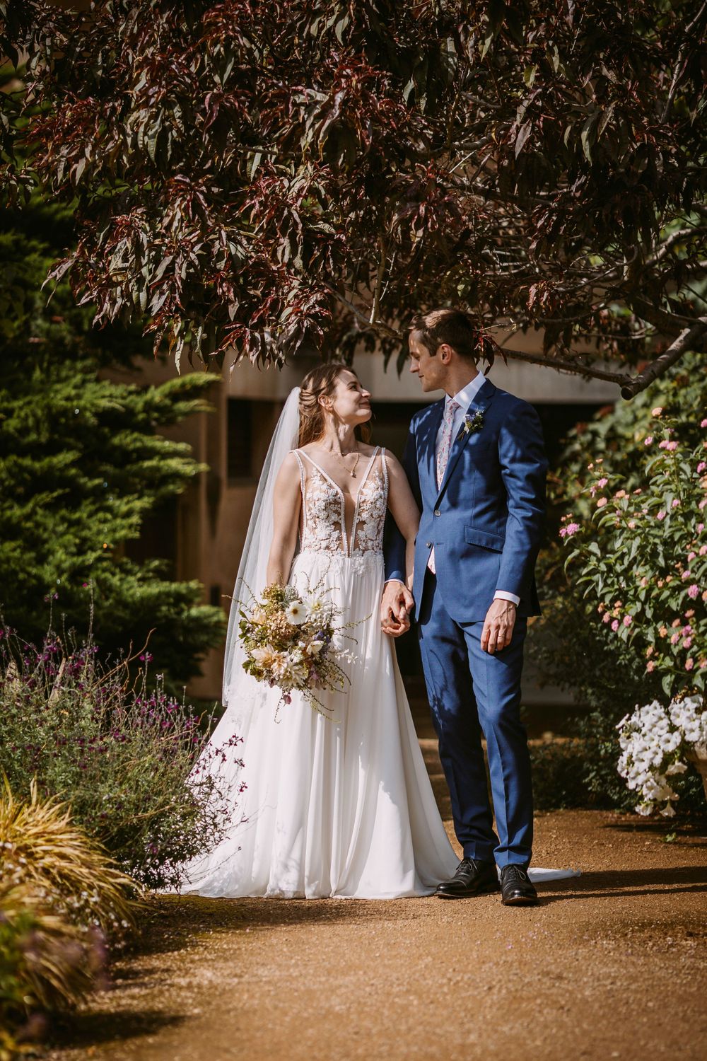 A wedding couple stands amid autumn foliage on a garden path, with the bride in white and groom in navy blue.