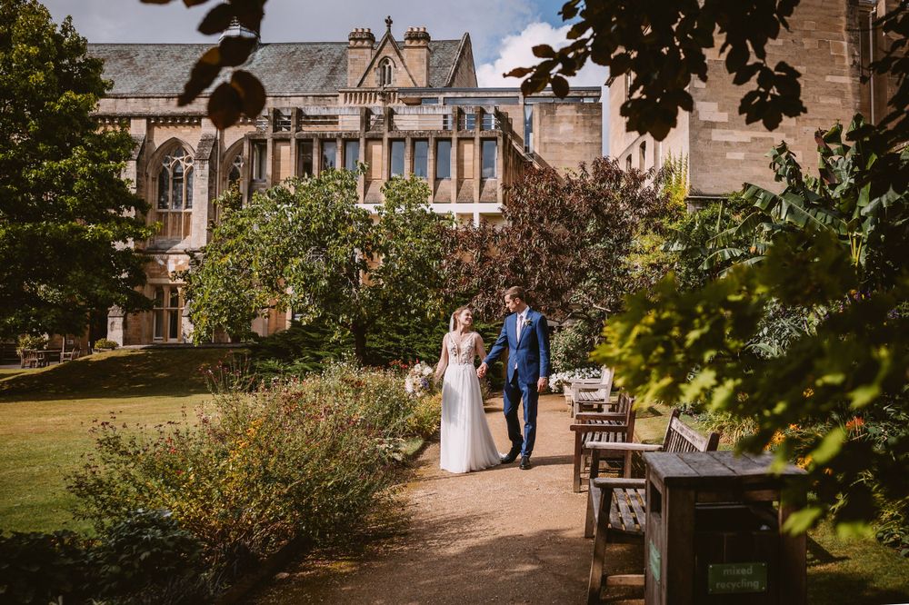 Couple walks along garden path beside historic stone building on sunny day at Balliol College, Oxford