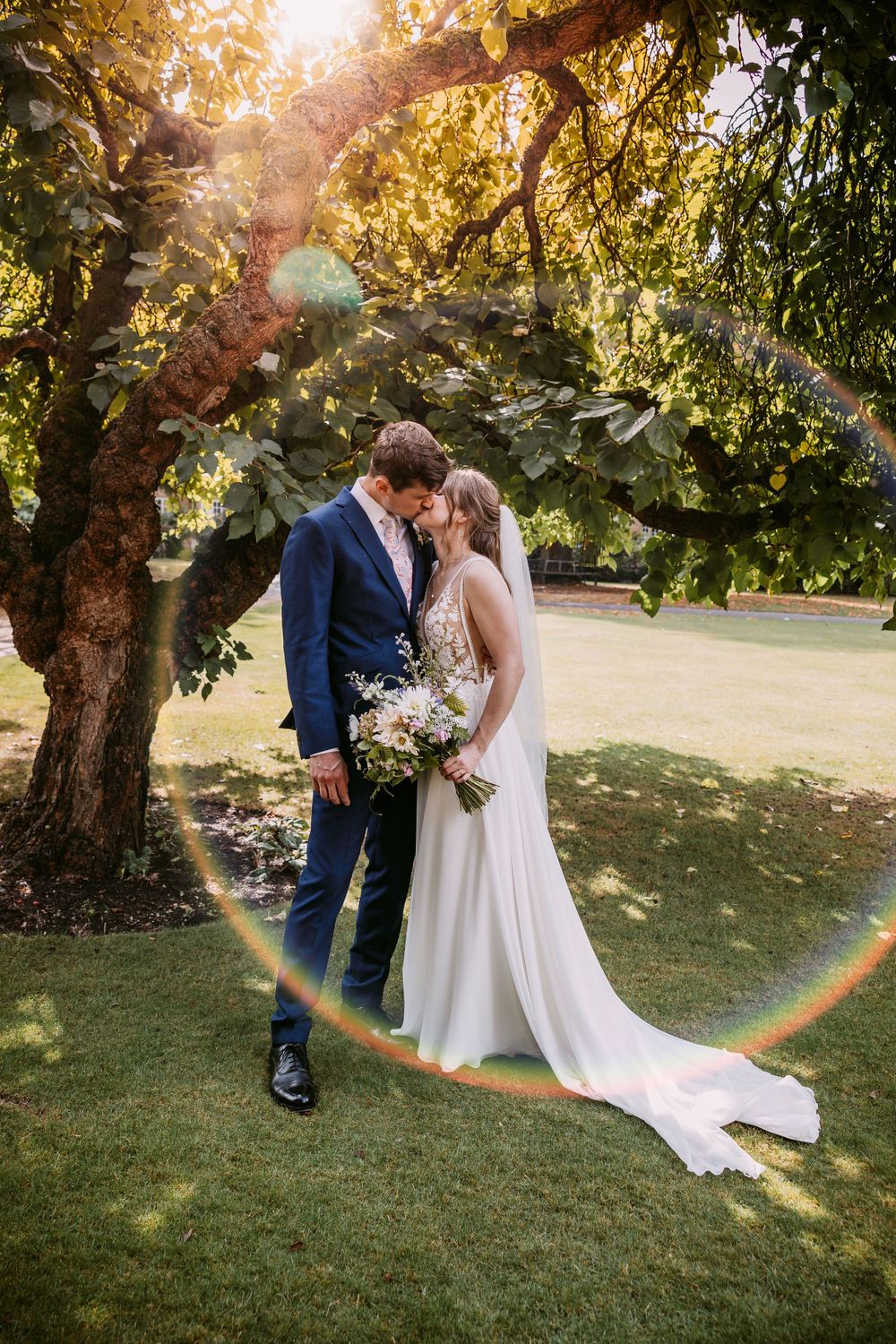 A romantic wedding portrait under a sunlit tree with a golden lens flare creating a circular effect.