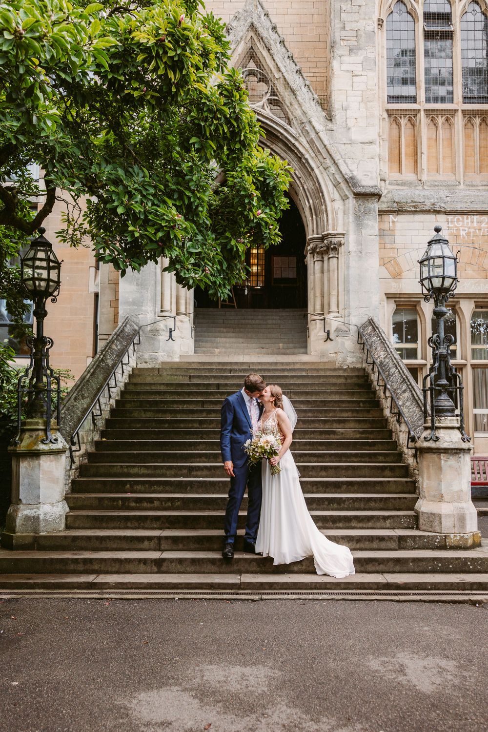 A wedding couple poses on grand stone steps leading to a the Grand Hall entrance at Balliol College, Oxford