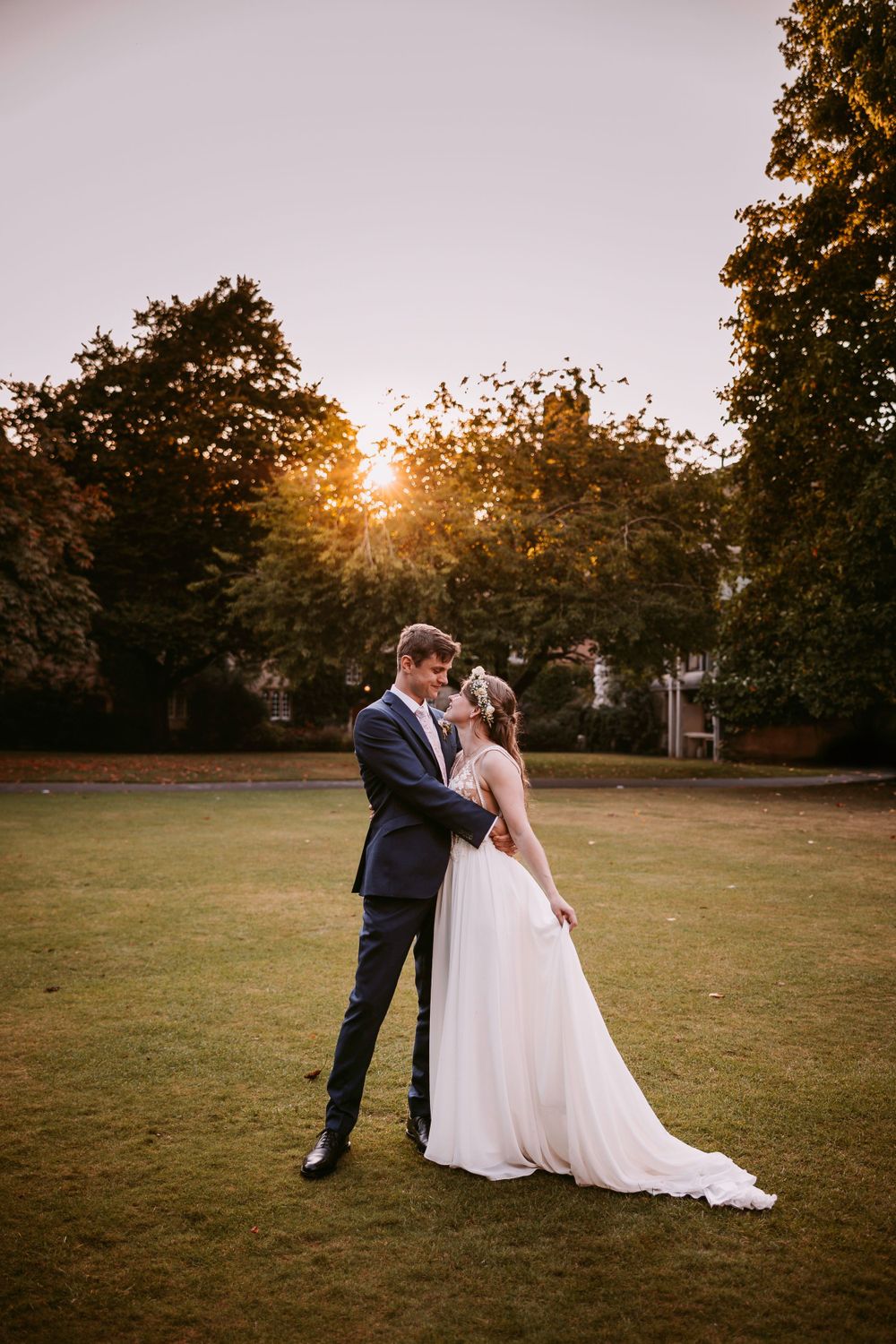 A romantic sunset wedding portrait on expansive lawn with golden light filtering through trees.