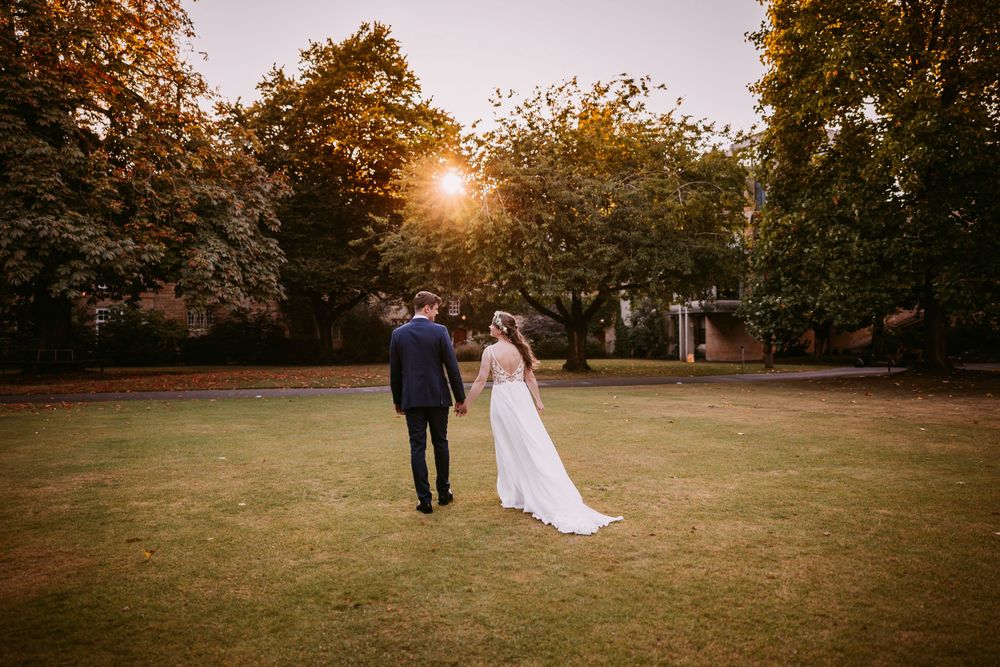 A wedding couple walks hand in hand across a lawn during golden hour with trees silhouetted against the sky at Balliol College, Oxford