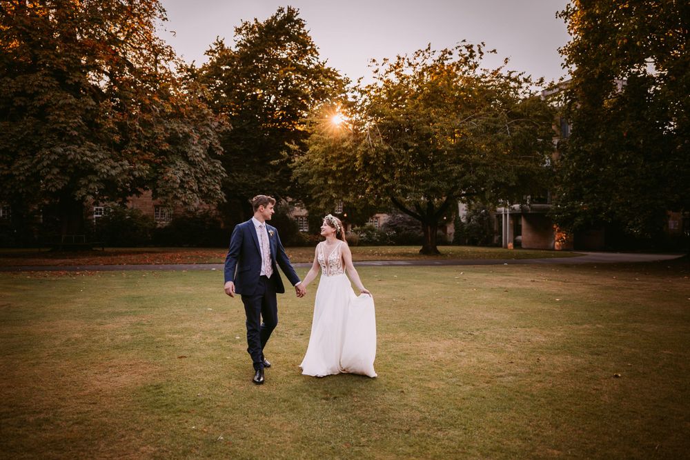 A couple in wedding attire walks together across an autumn lawn at golden hour at Balliol College, Oxford