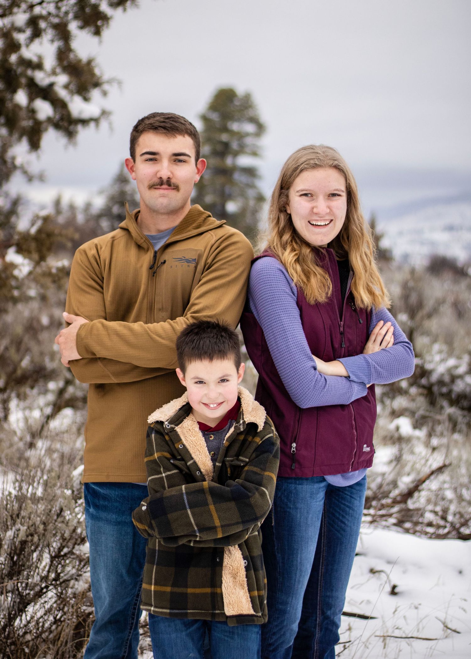 3 siblings stand together, the taller ones back to back with the younger just in front of them, all have crossed arms and smiling at camera