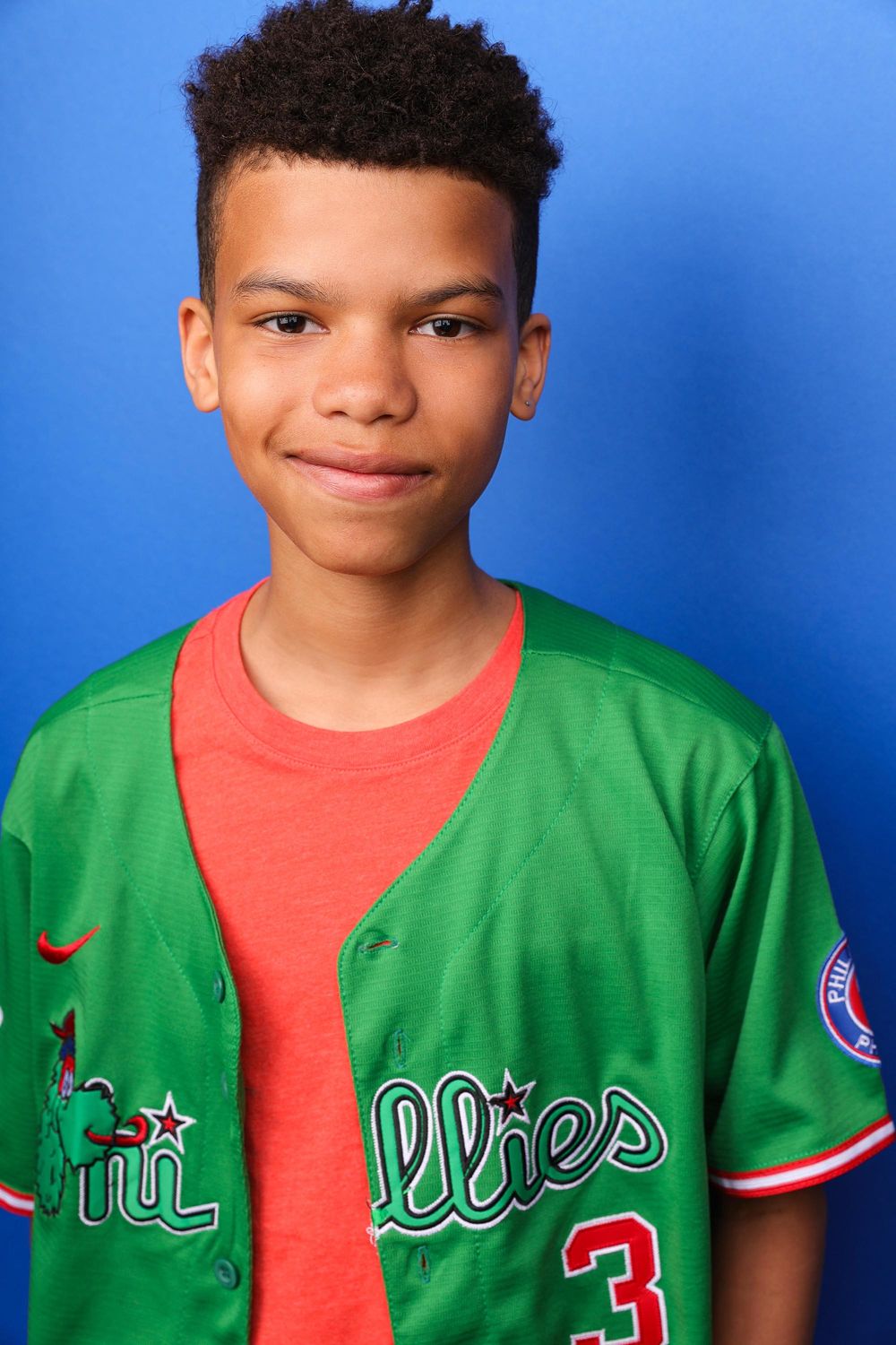 Young boy smiling in a green Philadelphia Phillies baseball jersey over a red t-shirt against a blue background.