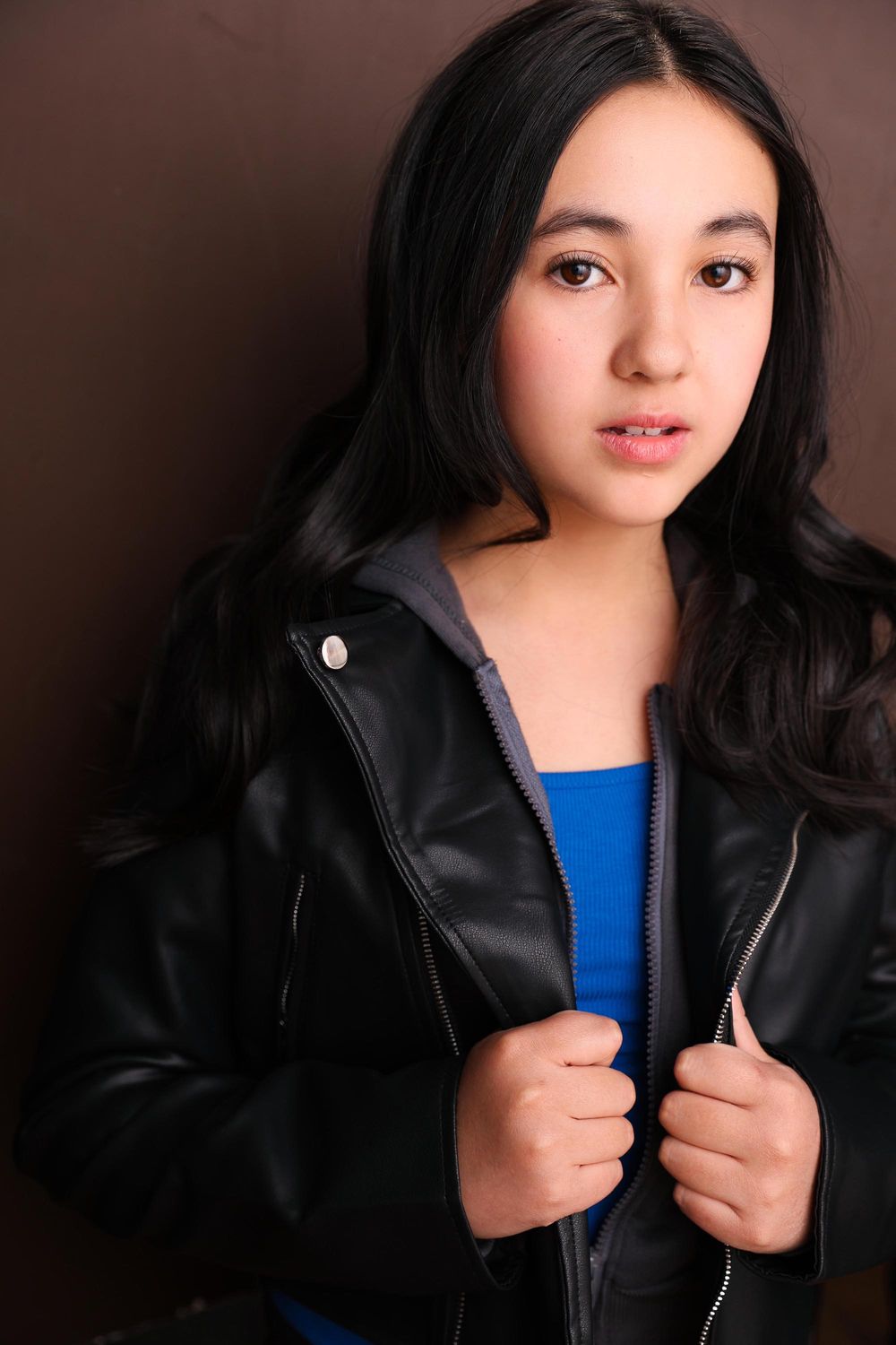 Young girl with long dark hair wearing a black leather jacket over a blue top, posing confidently in a studio.