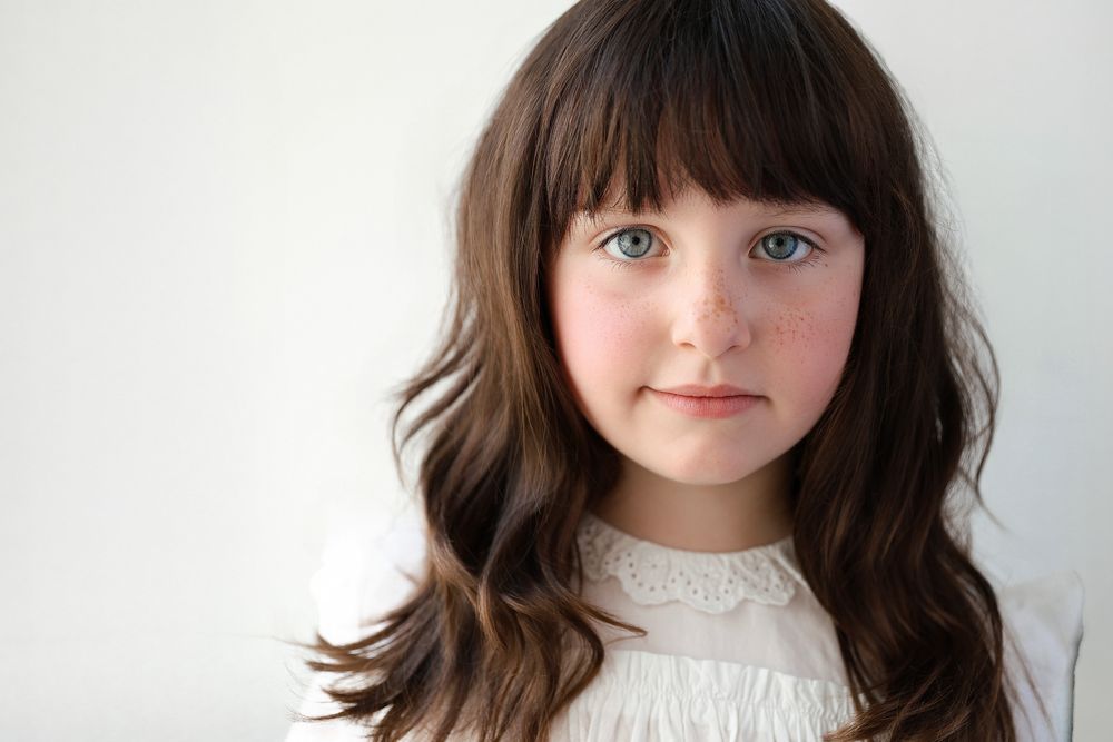 Young girl with long brown wavy hair and bangs, blue eyes, wearing a white lace top, portrait photo.