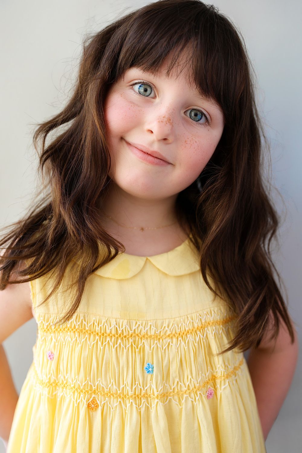 Young girl with brown hair and blue eyes wearing a yellow smocked dress, smiling softly at the camera.
