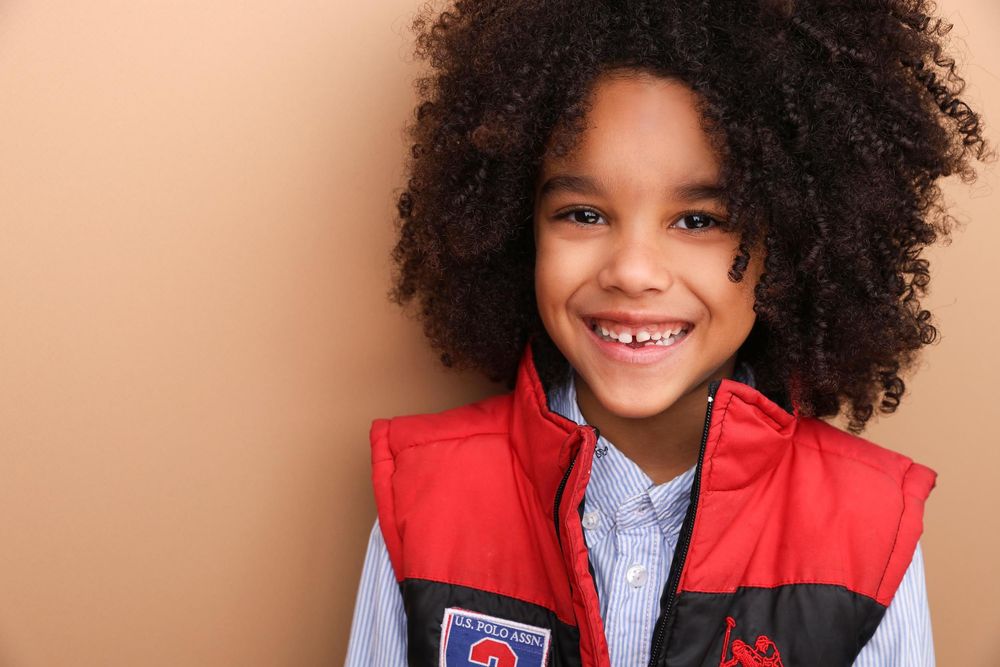 Smiling young boy with curly hair wearing a red vest and blue shirt against a beige background.