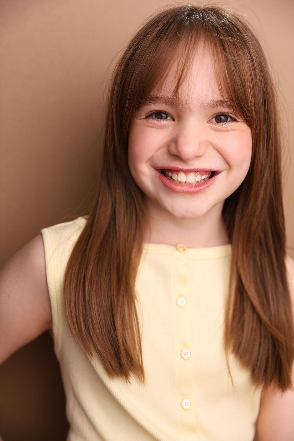 Smiling young girl with long brown hair wearing a yellow sleeveless top against a tan background.