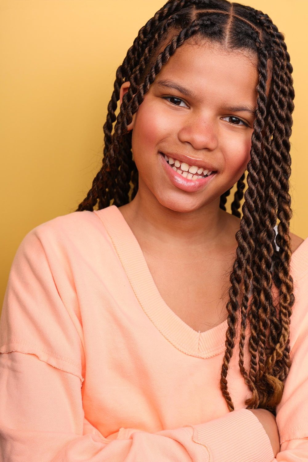 Smiling young girl with braided hair wearing a peach sweatshirt against a yellow background.