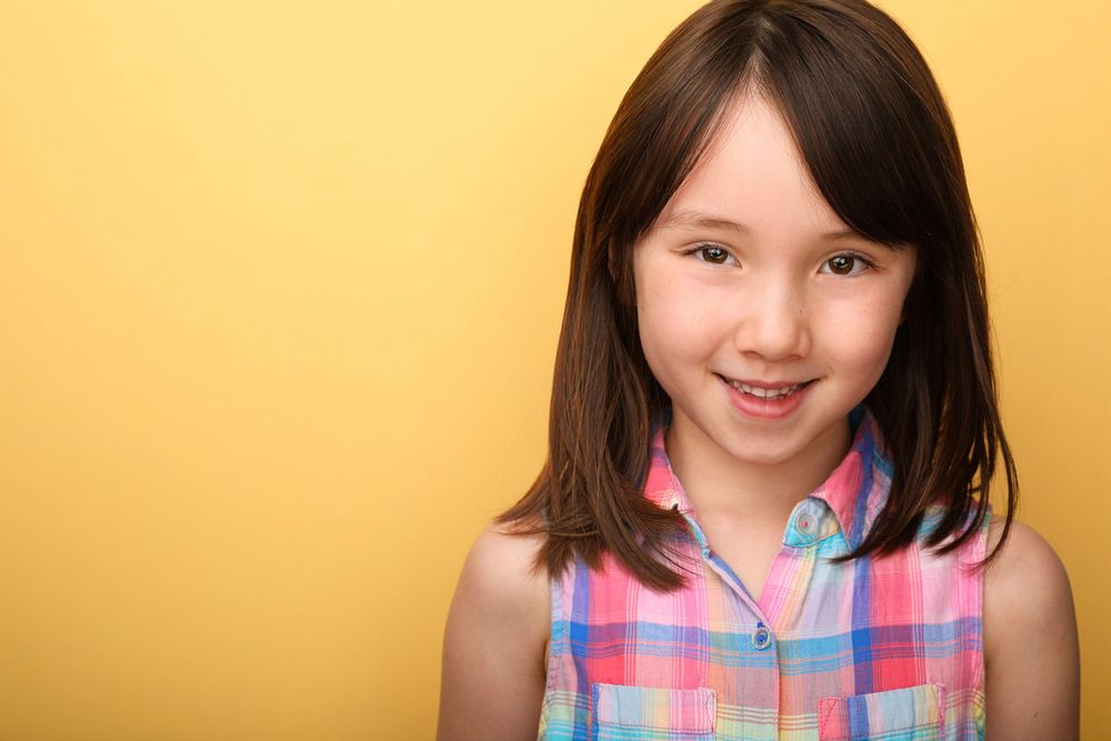 Young person in a colorful plaid sleeveless shirt smiling against a warm yellow background.