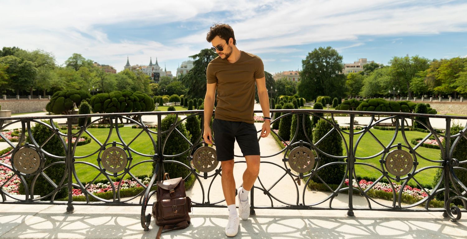 Person standing by an ornate railing overlooking a scenic park on a sunny day with a bag nearby.