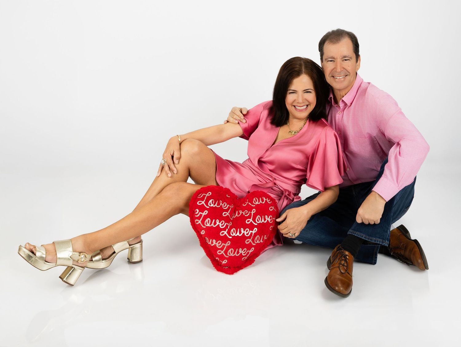 Studio portrait with couple in pink and casual attire posing with heart-shaped prop against white background.