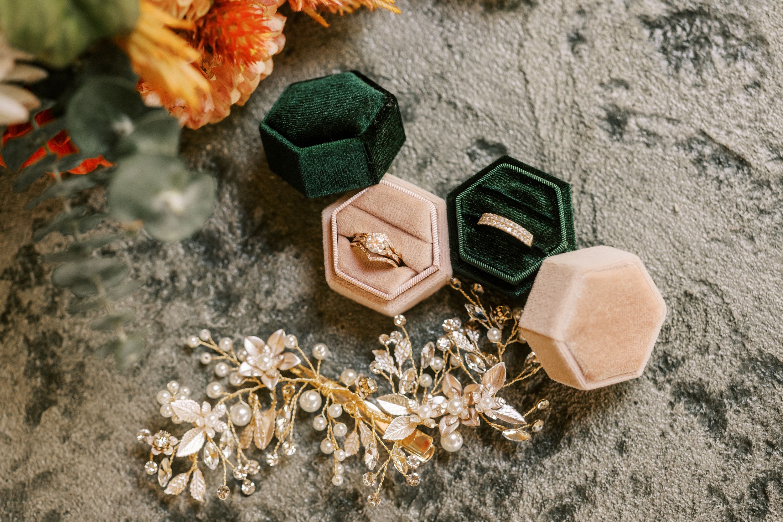 Wedding details including velvet ring boxes and floral hair accessories on textured surface.