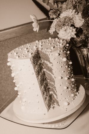A white wedding cake with pearl decorations and a cut slice showing layered interior next to fresh flowers.