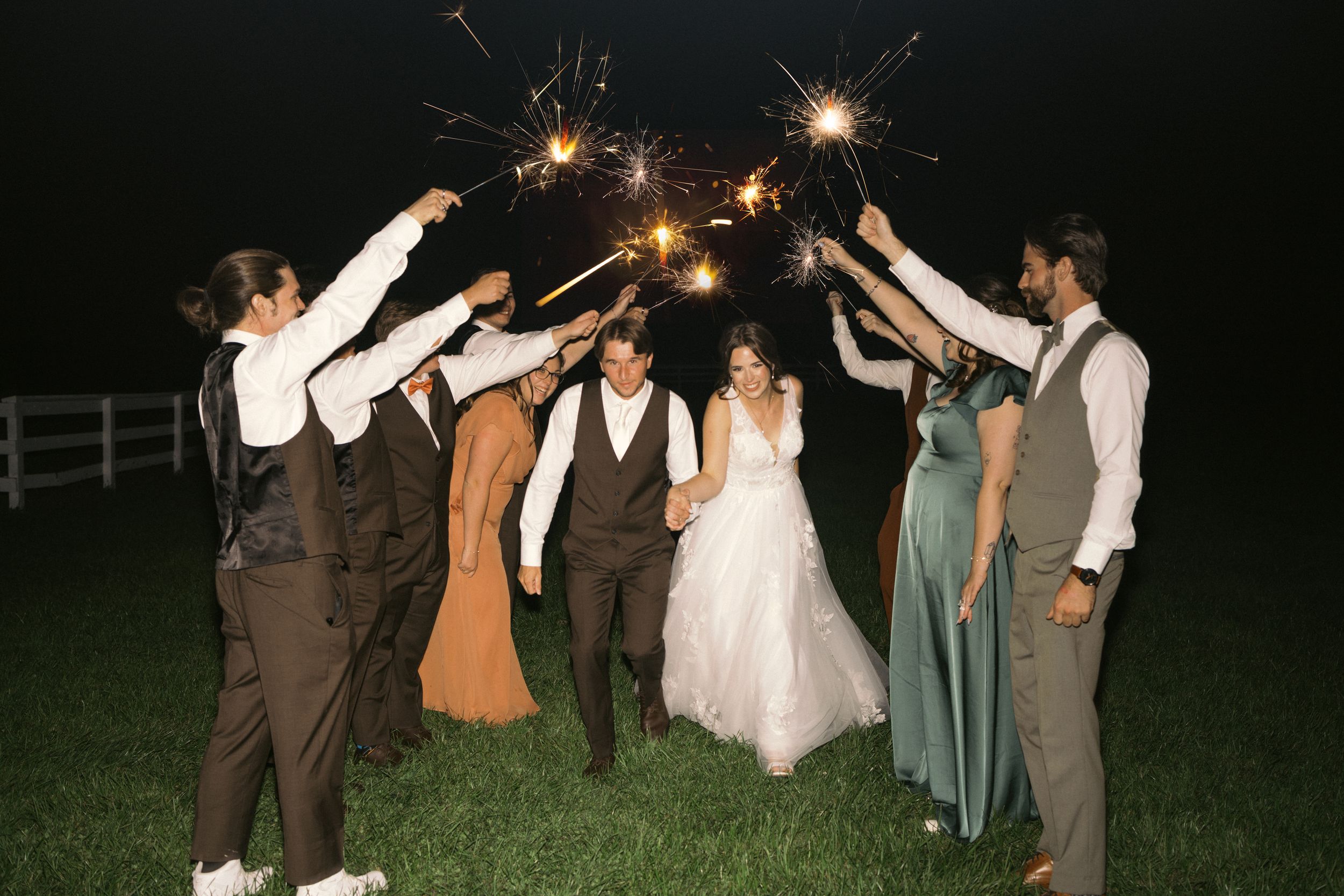 Wedding party holds sparklers in celebration on a grassy field at night.