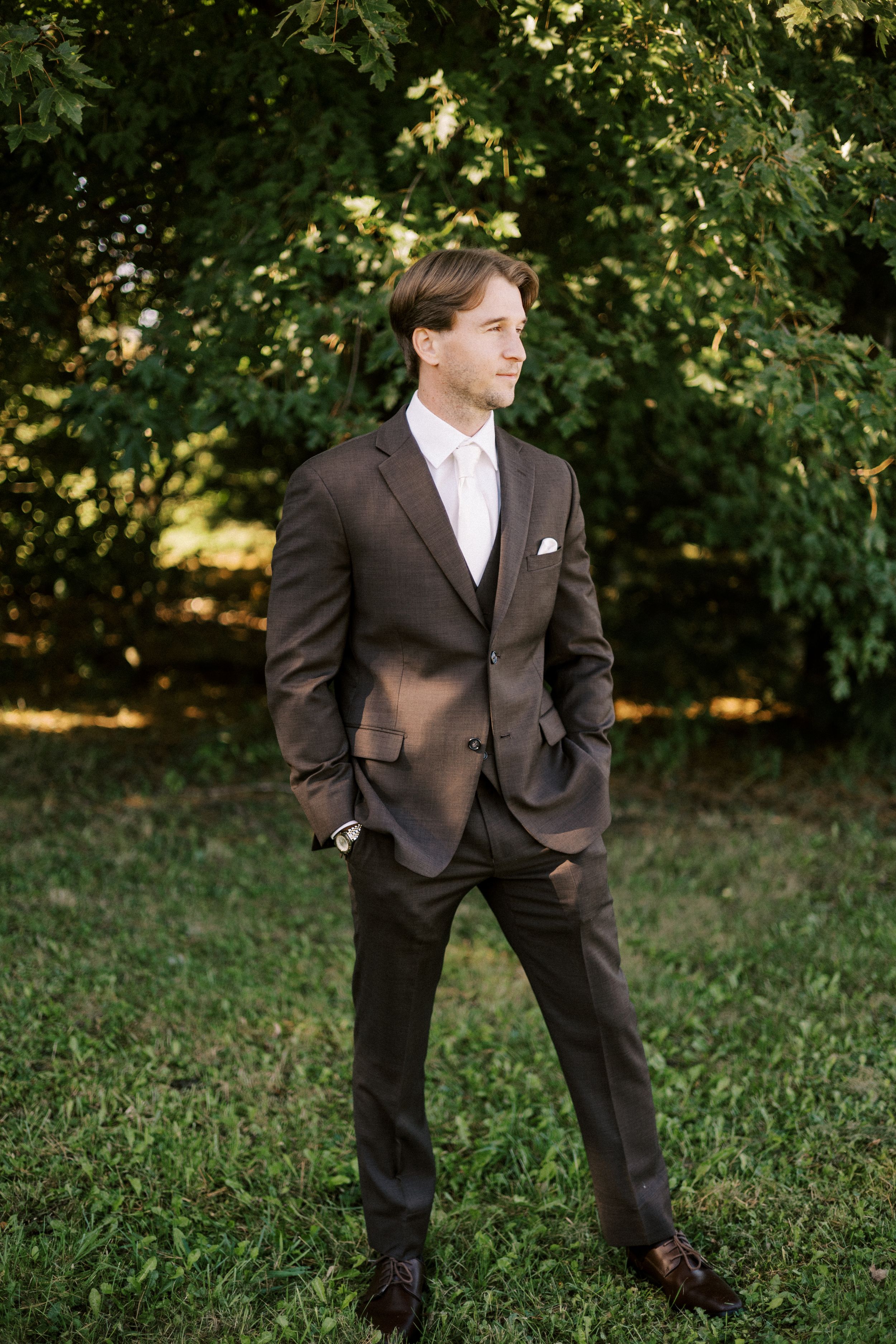 An elegant groom in a brown suit poses for a portrait in a garden setting.