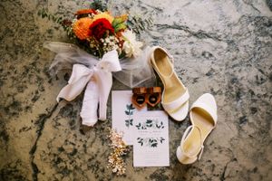 Wedding details flatlay with white shoes, florals and invitation on marble surface.