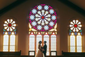 A couple embraces in front of a stunning stained glass church window with snowflake patterns and warm light streaming through.
