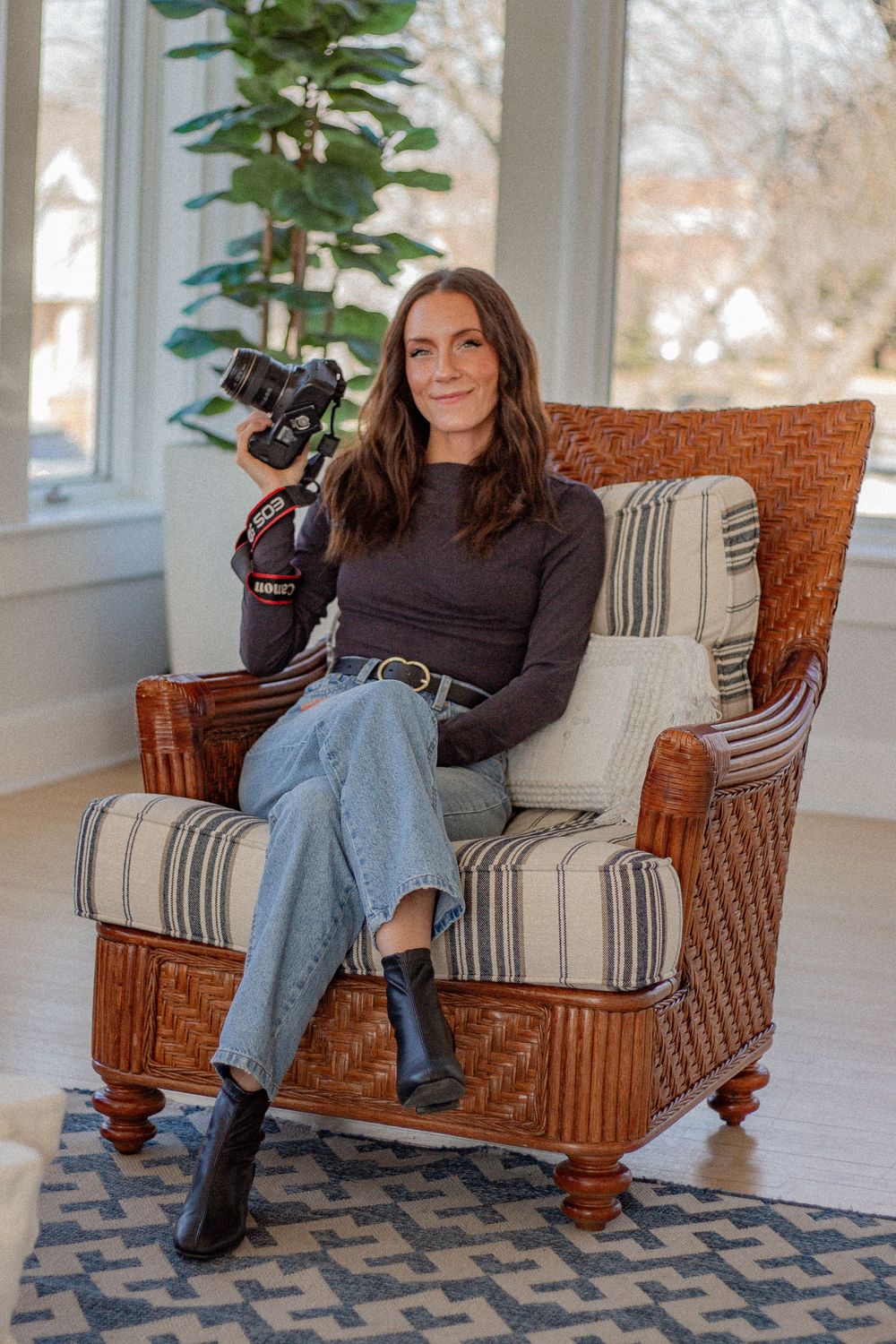Woman sitting in wicker chair holding a camera, wearing jeans and dark top, in a bright sunlit room.