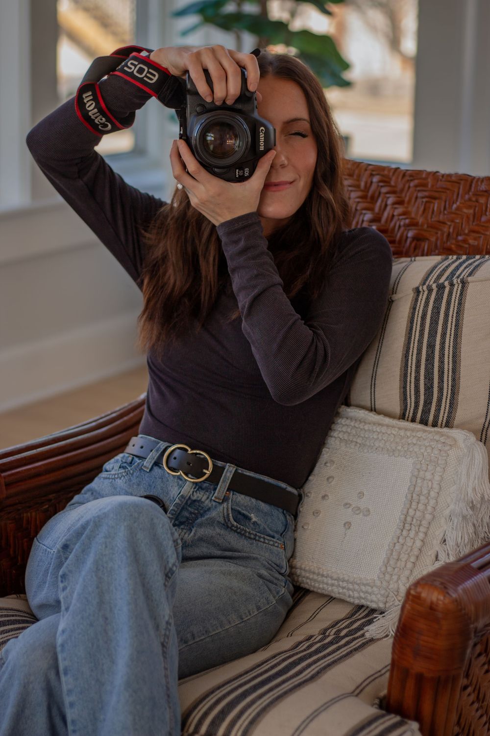 Woman sitting in wicker chair holding Canon EOS camera up to take a photo, wearing jeans and dark turtleneck.