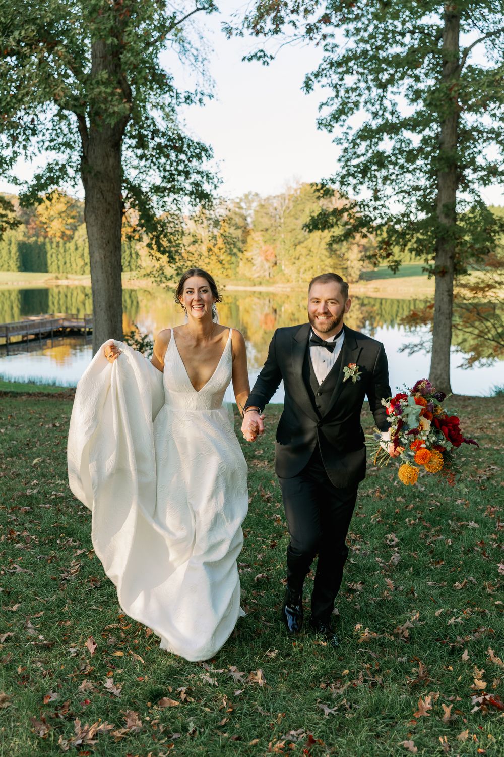 A couple runs playfully through fallen leaves on their wedding day surrounded by autumn trees.