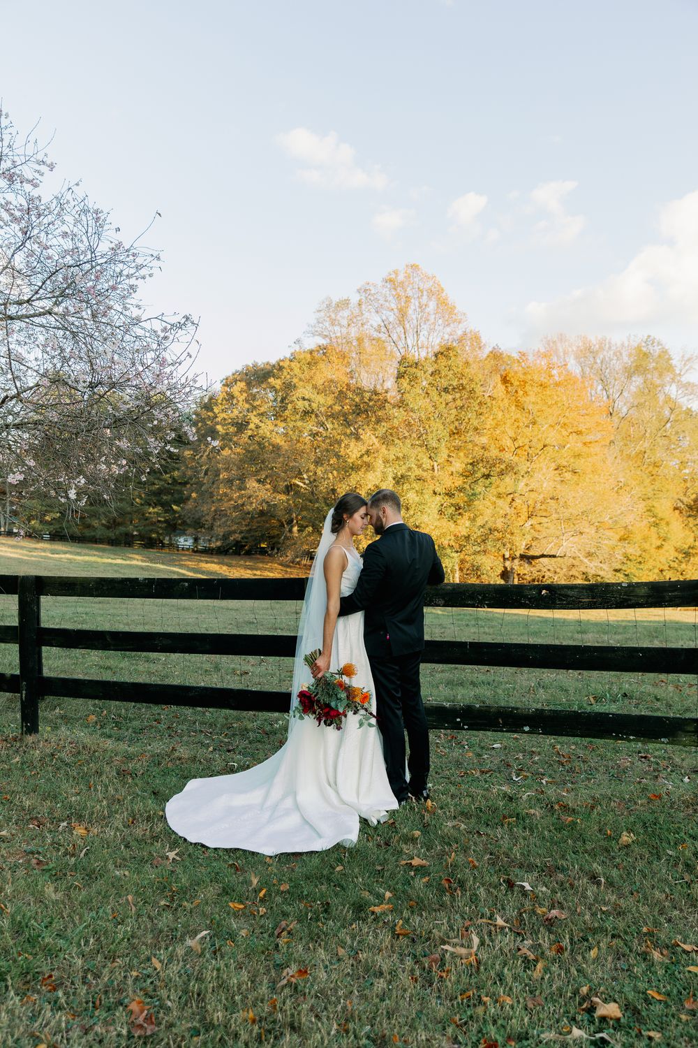 A couple poses together on grassy hillside with autumn foliage and wooden fence in background.